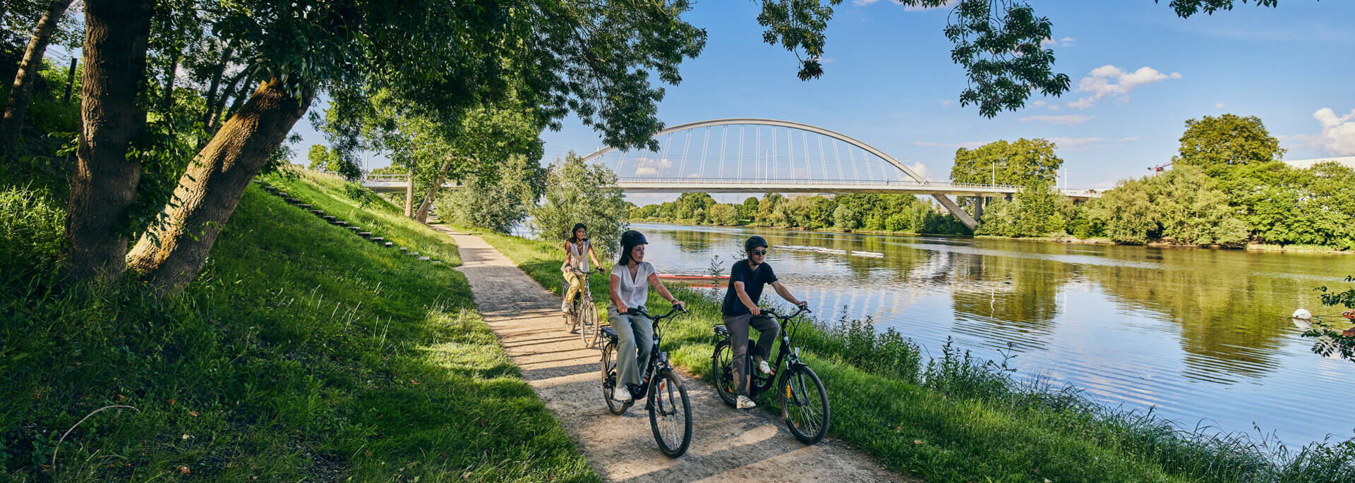 3 young people cycling along the waterfront, Confluences Bridge in the background.
