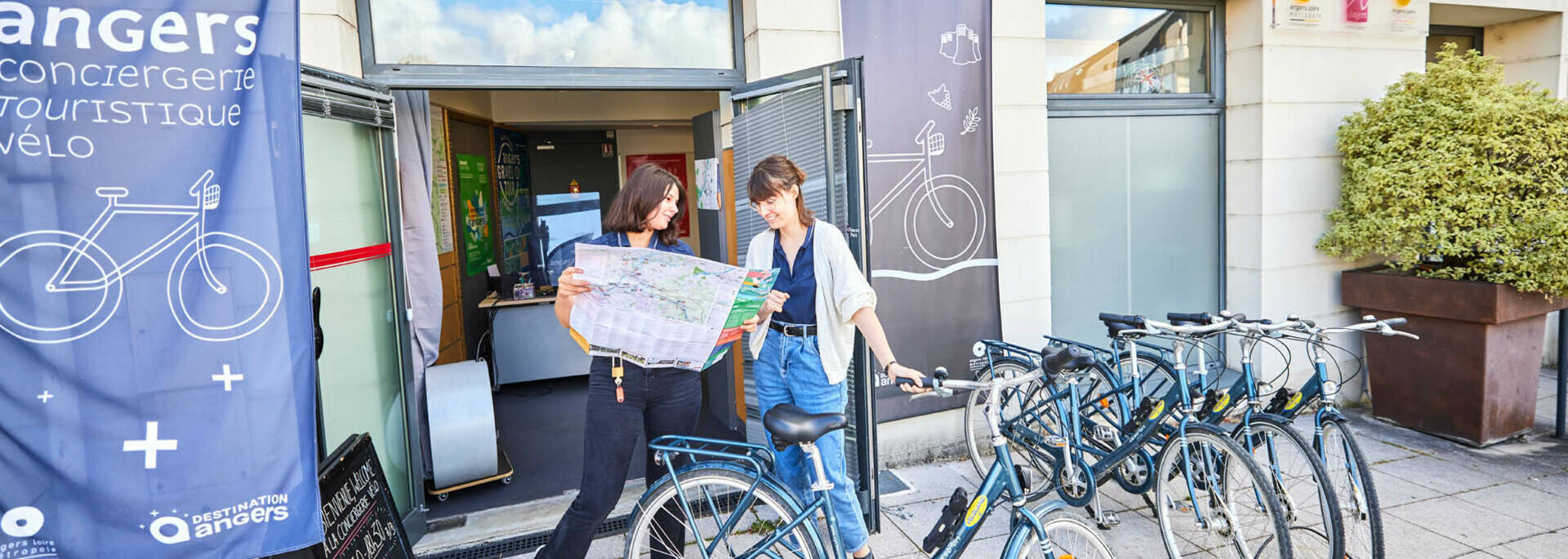 Deux femmes regardant un plan devant la Conciergerie vélo de l'Office de tourisme d'Angers, nombreux vélos rangés devant