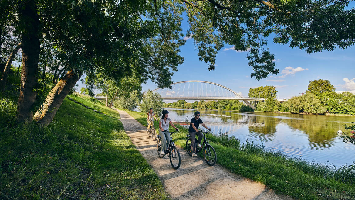 3 young people cycling along the waterfront, Confluences Bridge in the background.