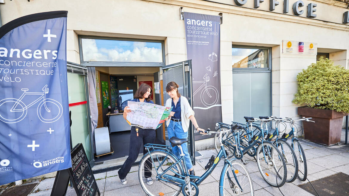 Deux femmes regardant un plan devant la Conciergerie vélo de l'Office de tourisme d'Angers, nombreux vélos rangés devant