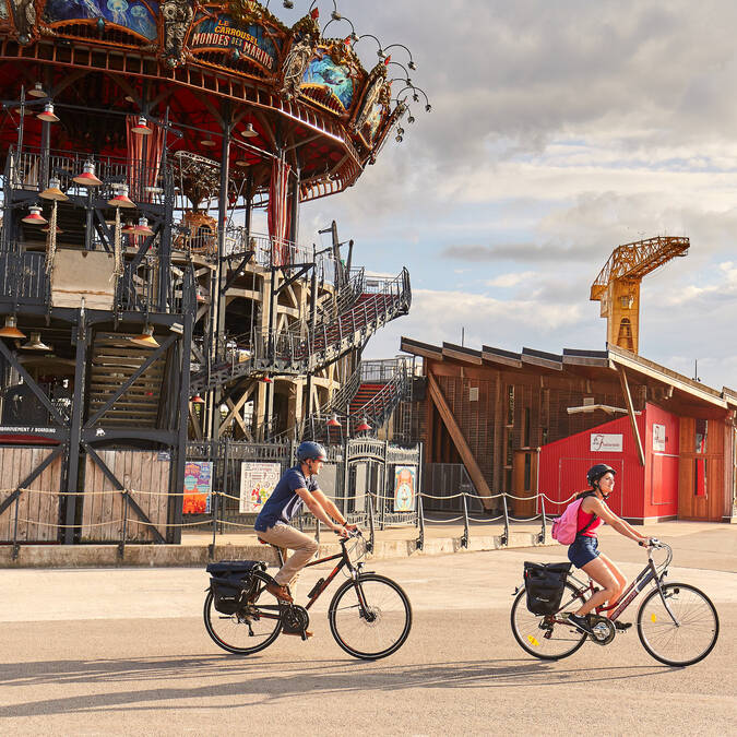 Couple on bicycles passing in front of the Carrousel of the Marine Worlds, the yellow crane in the background